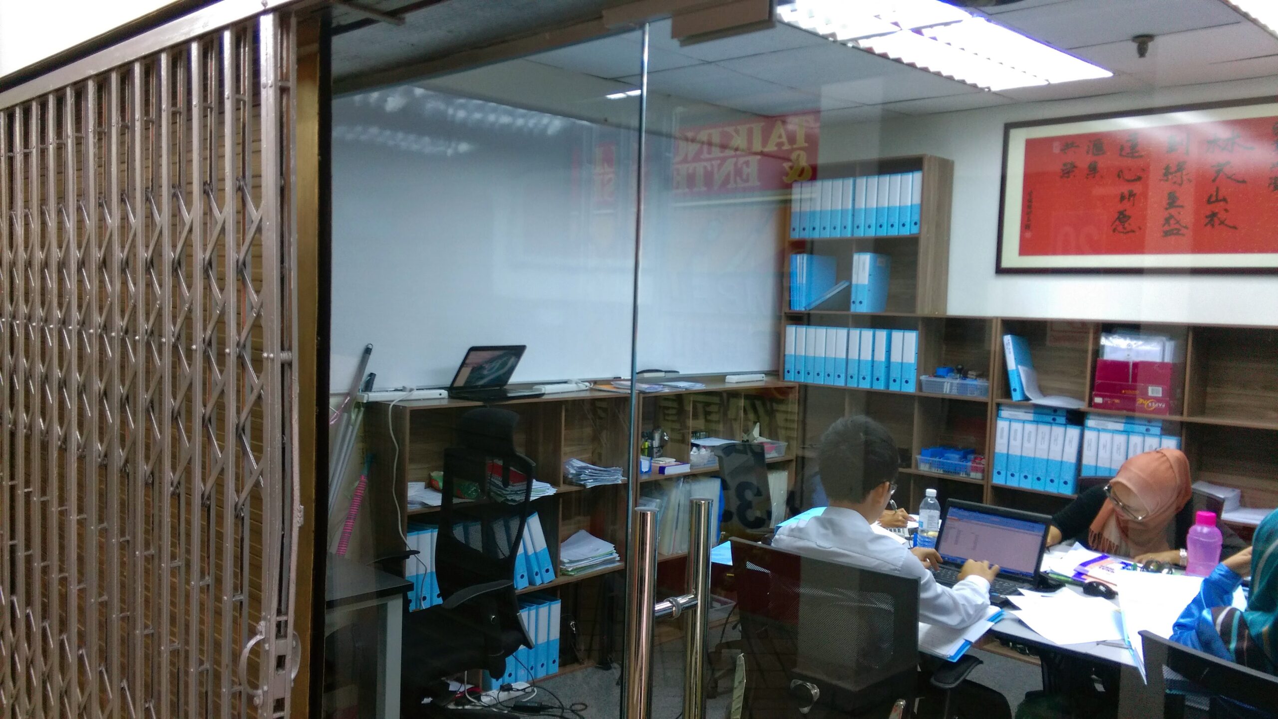 Interior view of a small, well-organised office with glass walls and a closed metal shutter gate. Two team members are seated at desks, working on laptops surrounded by shelves stacked with blue client files.